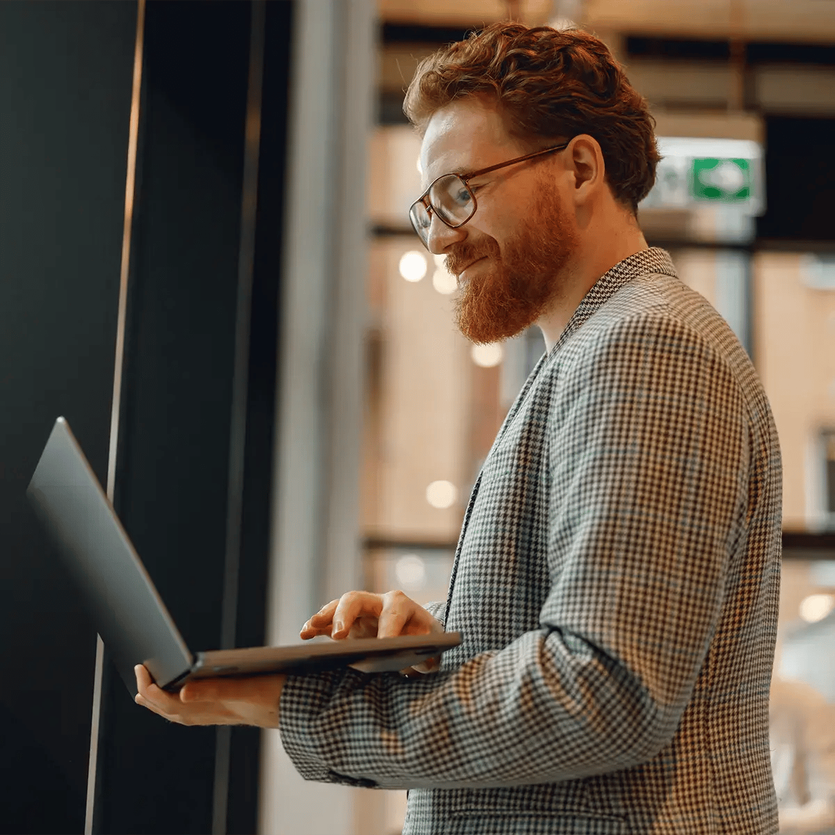 A bearded man in a checkered blazer smiles while using a laptop