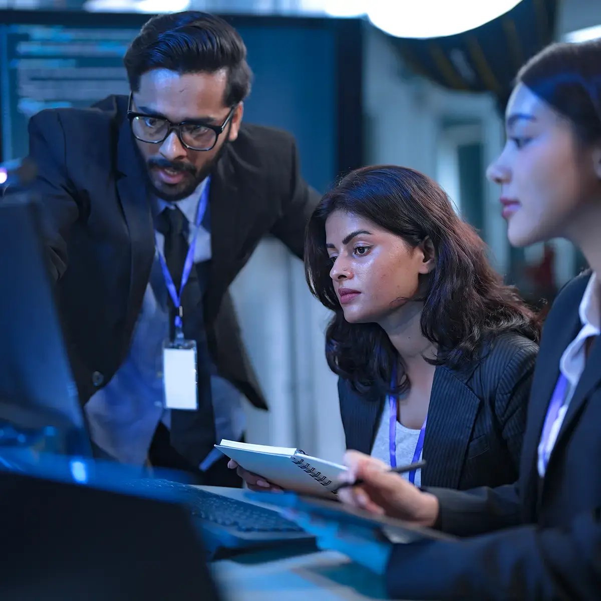 A diverse team of three professionals in formal attire focuses intently on computer screens in a dimly lit office