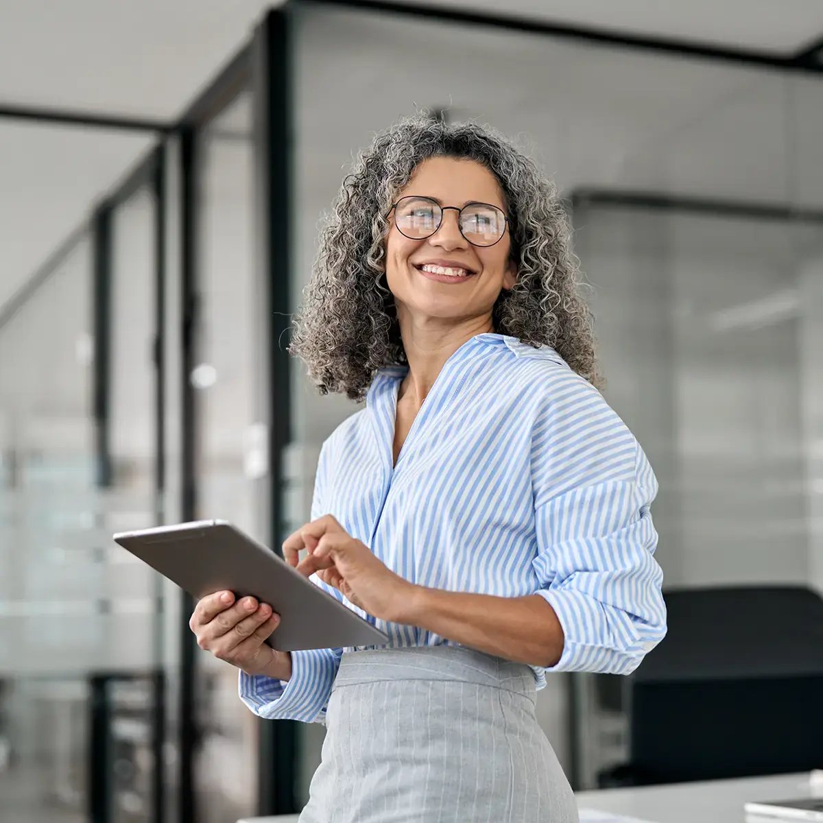 A smiling woman with curly gray hair and glasses holds a tablet in a modern office