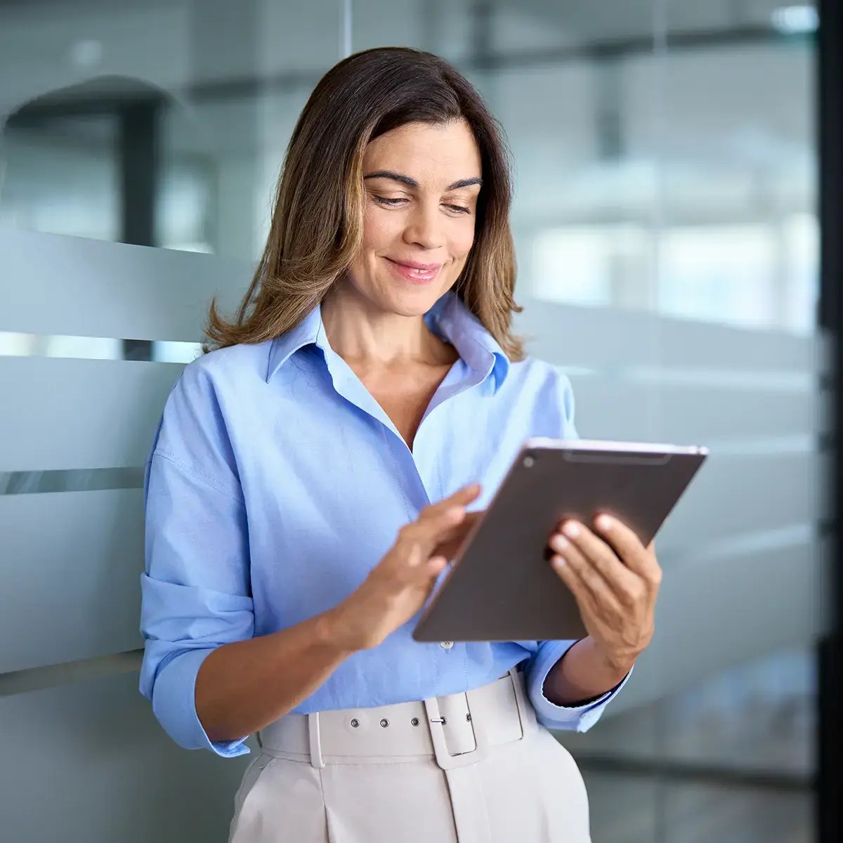 A woman in a light blue blouse smiles while using a tablet, standing in a modern office with glass walls