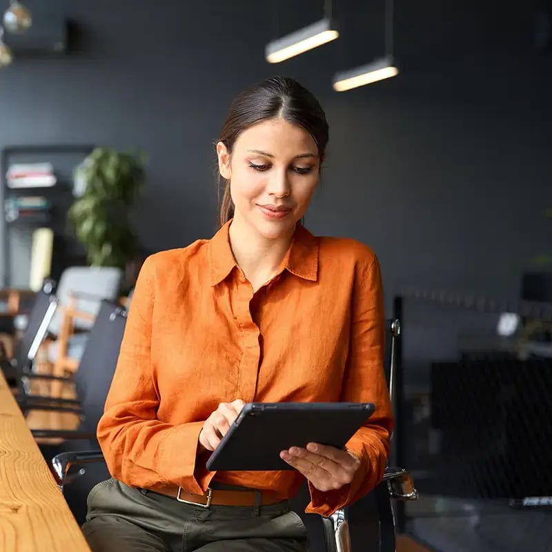 A woman in an orange blouse sits at a wooden desk in a modern office, using a tablet