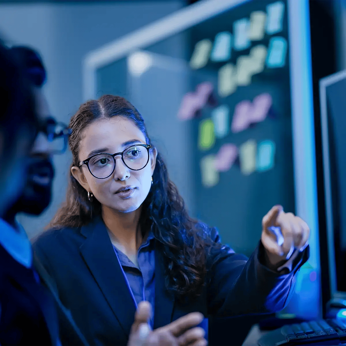 A woman in glasses gestures while discussing work with colleagues at a computer
