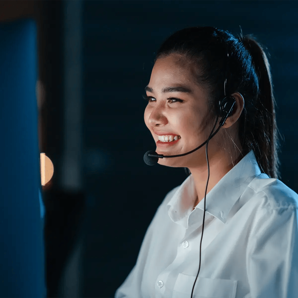 A woman wearing a headset smiles while working at a computer in a dimly lit room