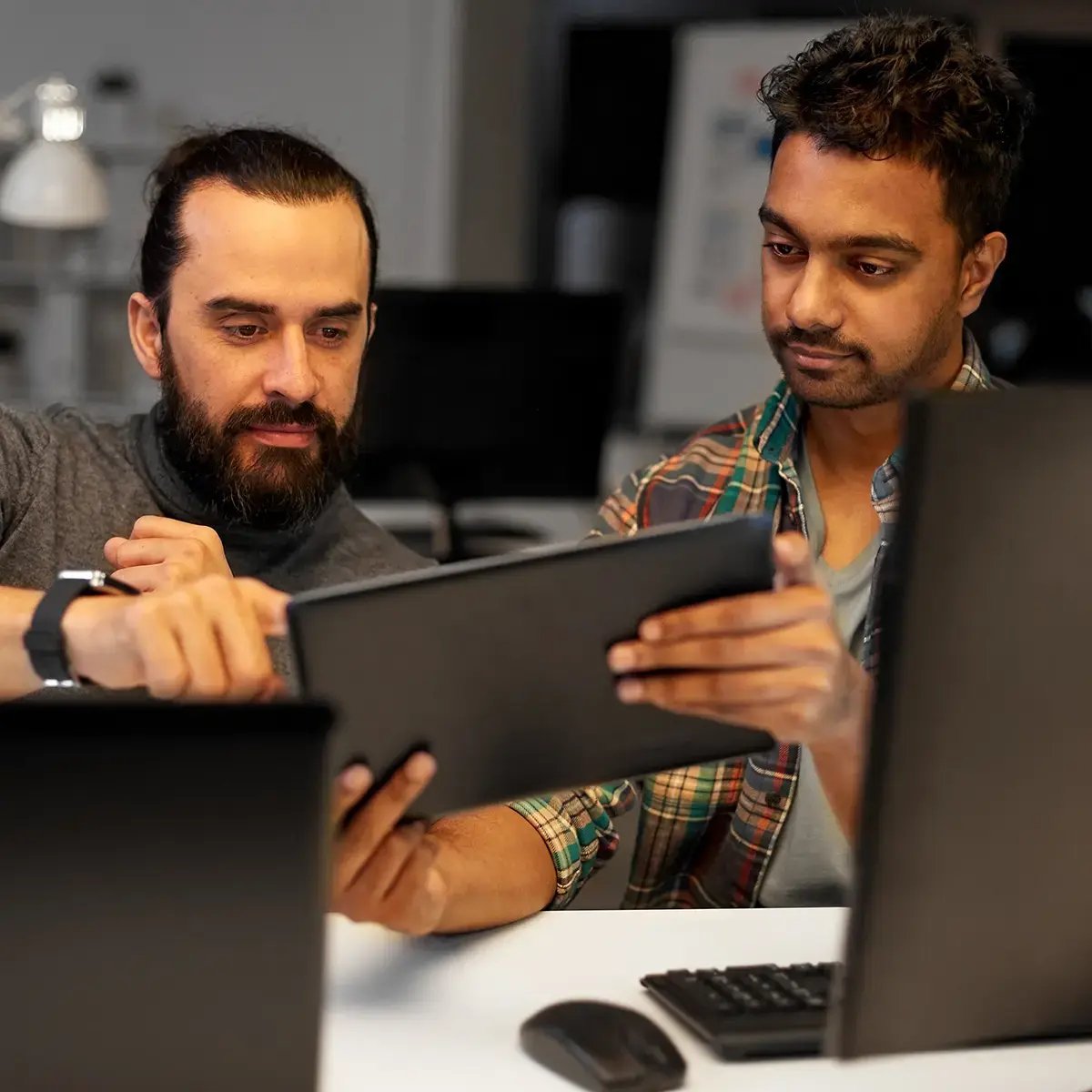Two men in an office setting collaborate over a tablet, with focused expressions