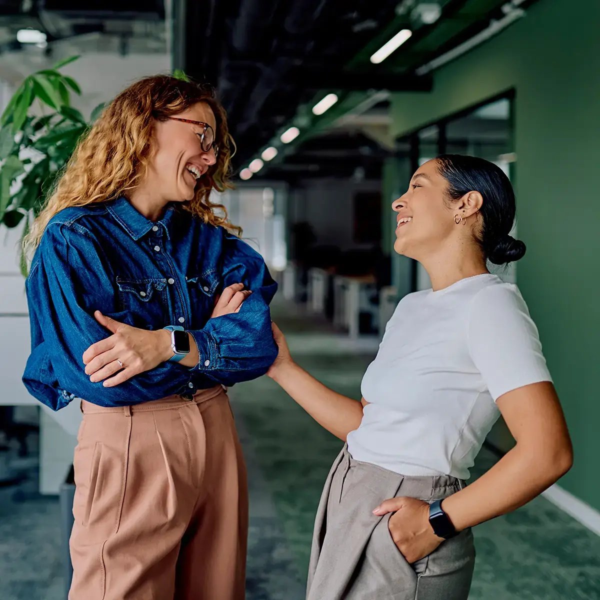 Two women are laughing and talking in a modern office hallway