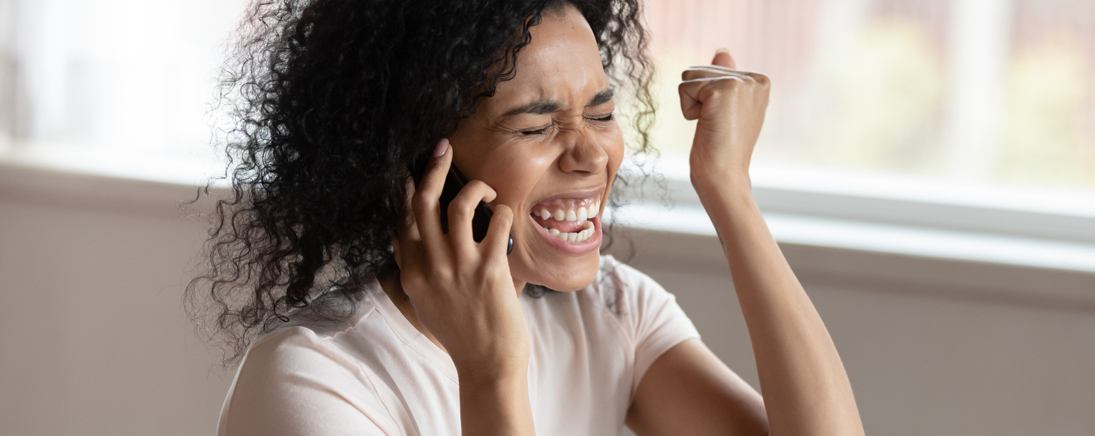 Overjoyed euphoric happy black woman sitting at desk talking on smartphone