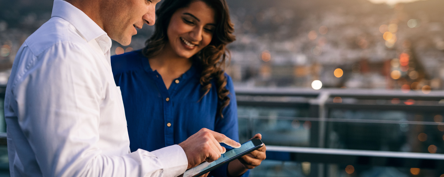 Two smiling coworkers working with a digital tablet together while standing on an office building balcony overlooking the city at dusk