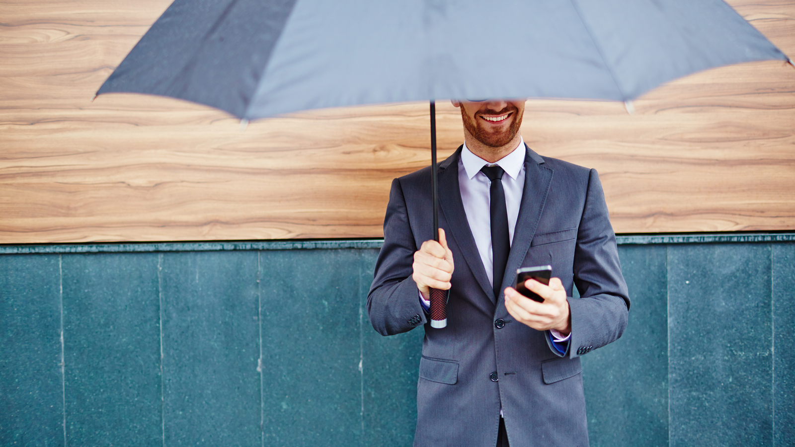 Happy young businessman with smartphone standing under umbrella