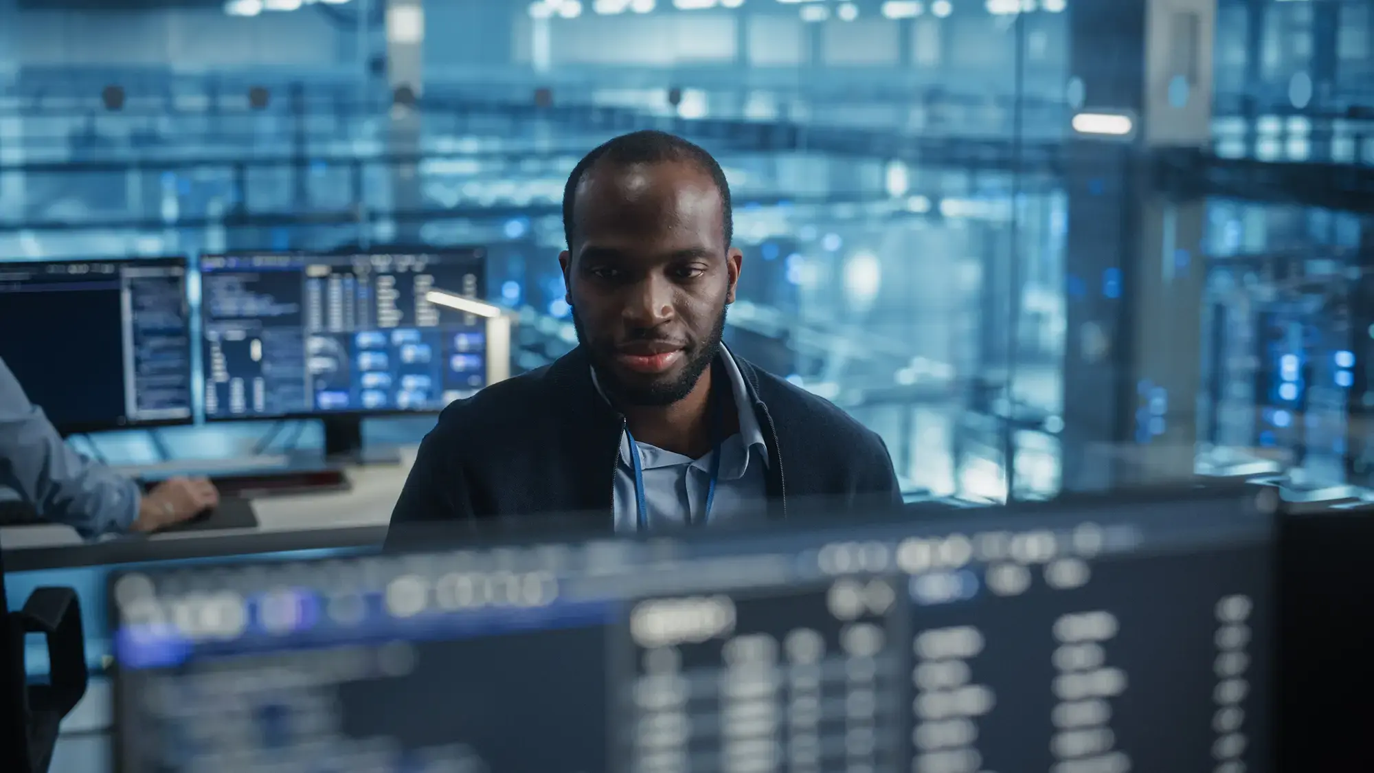 A man in a blue shirt focuses intently on multiple computer screens displaying complex data in a modern, glass-walled office or tech center