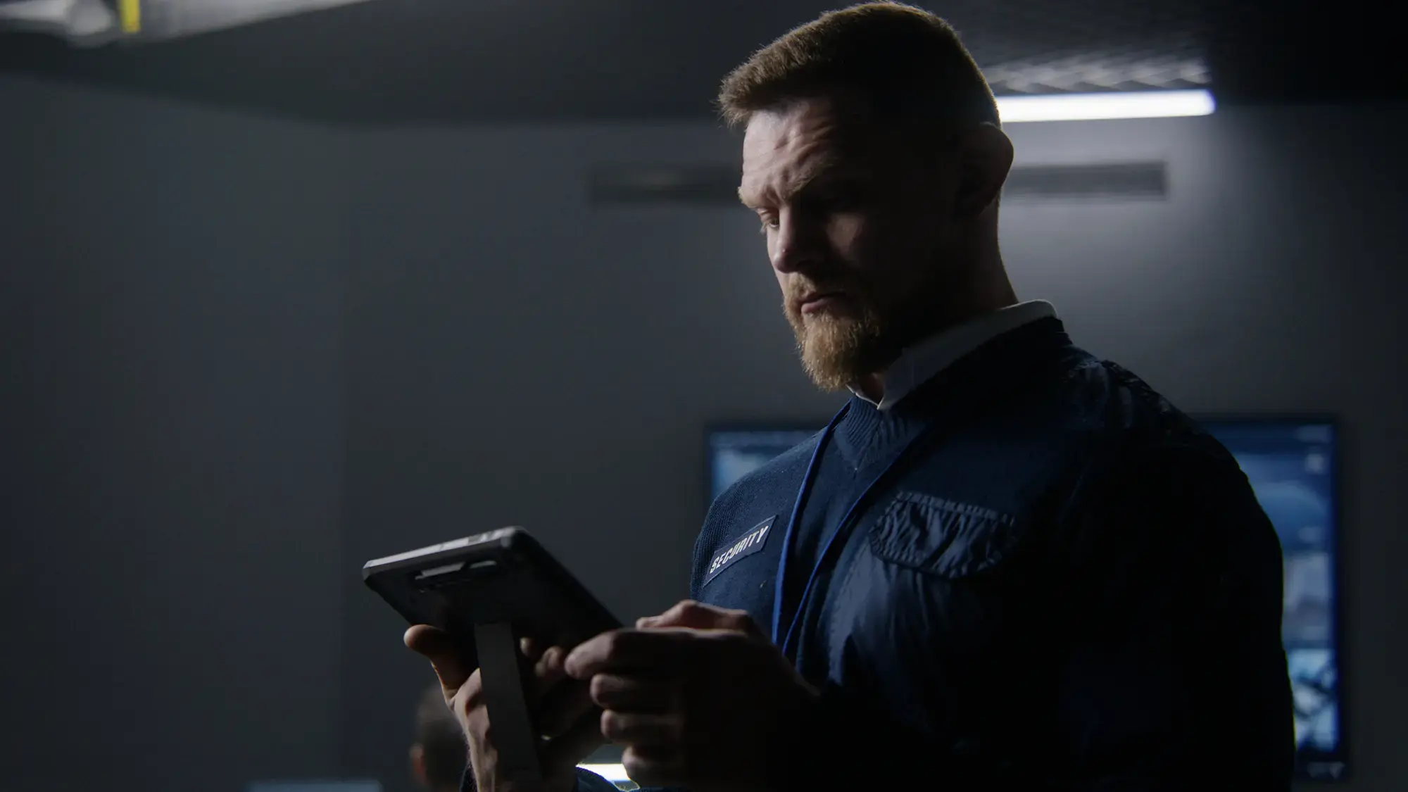 A man in a dark room studies a digital tablet with intense focus