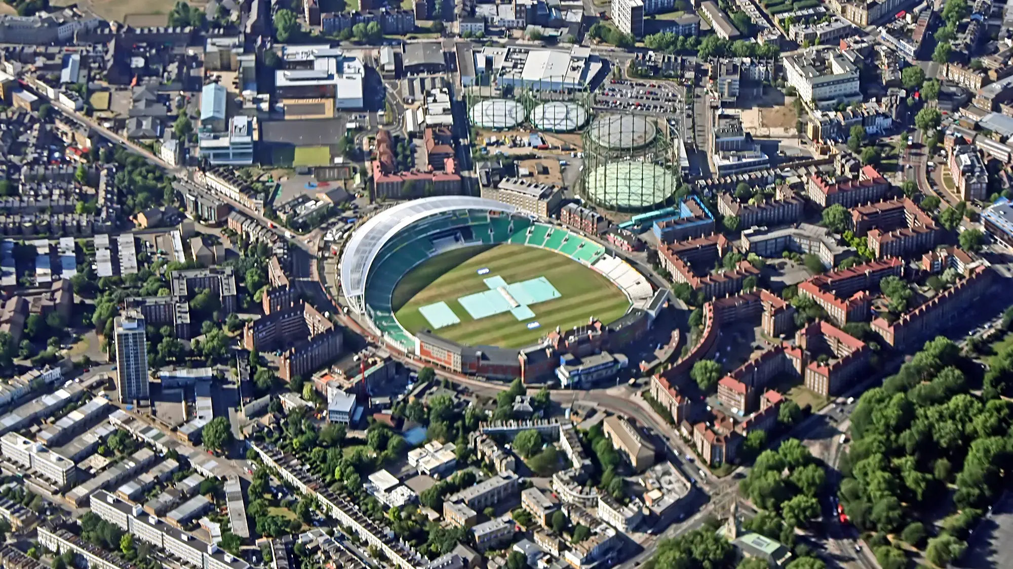 Aerial view of a large cricket stadium with a green field, surrounded by urban buildings and roads
