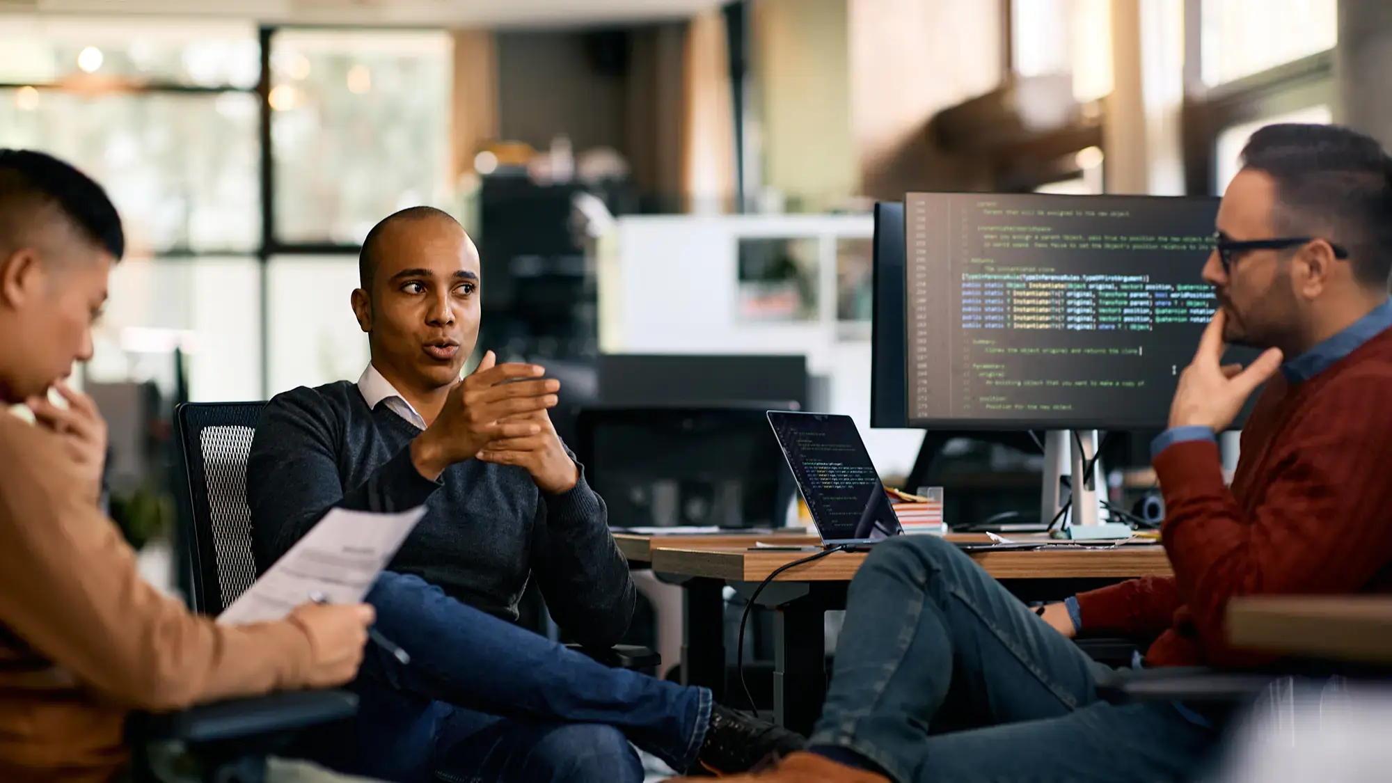 Three people in an office discuss ideas. A man gestures while talking, and others listen thoughtfully