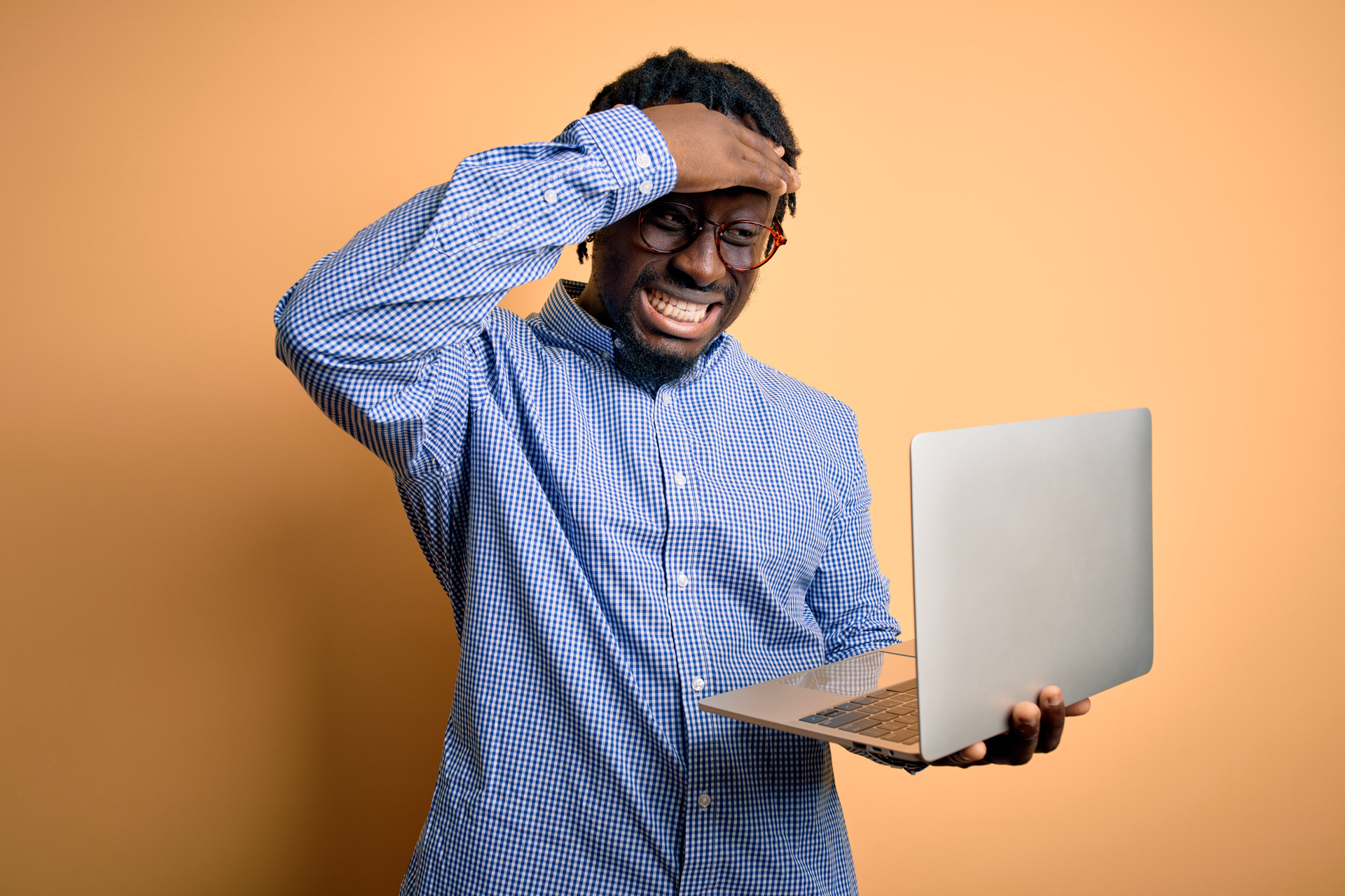 Young man using laptop standing in front of yellow background stressed with hand on head
